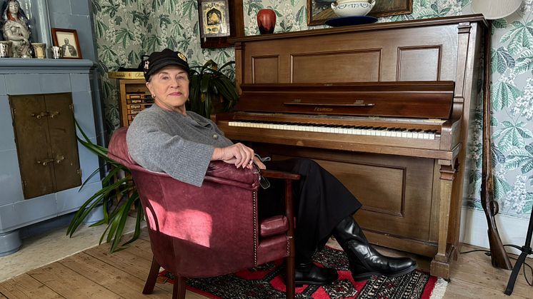 Py Bäckman sitter i en fåtölj framför ett piano i ett hemtrevligt rum med blommig tapet.