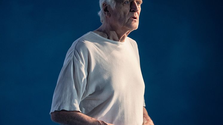 En äldre man tittar framför sig. Han bär vit t-shirt och bakgrunden är blå.