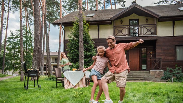 Familie im Garten vor einem Haus: Vater spielt mit Tochter Fußball, Mutter steht am gedeckten Tisch neben einem Grill.