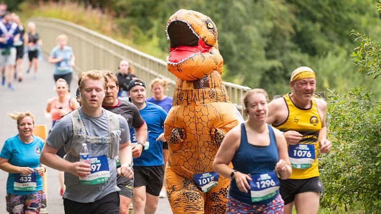 People running on a country tail in a long distance race with one participant wearing a dinosaur costume. 