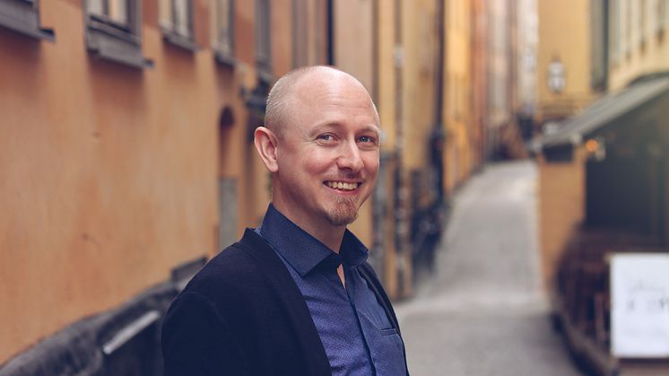 Danne ler stort, han har en kortklippt hår och lite skägg på hakan. Han har blå skjorta på sig, foto.
