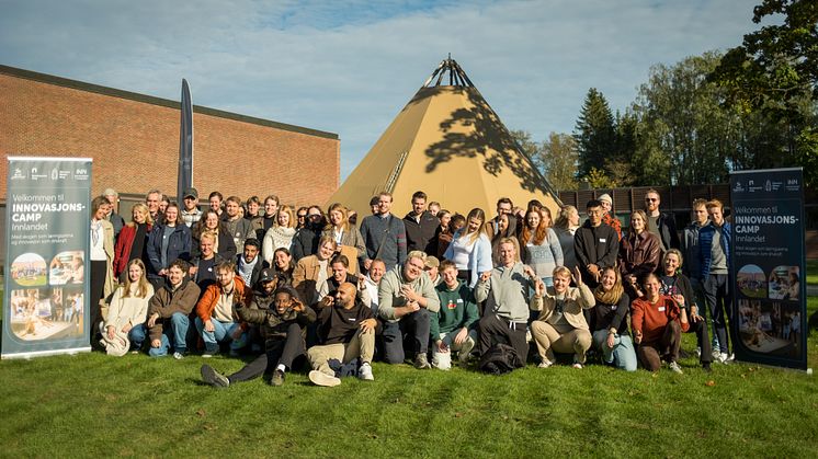 Stor gruppe studenter på gressplen foran en lavvo og mursteinsbygning