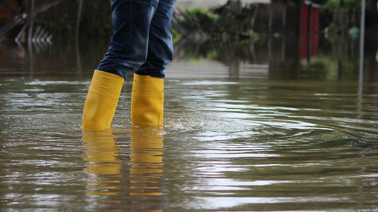 Person steht mit gelben Gummistiefeln in kniehohem Hochwasser auf einer überfluteten Straße.