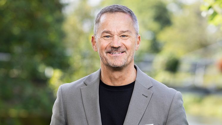 Bilden visar en leende person i grå kavaj och svart t-shirt mot grön bakgrund.
