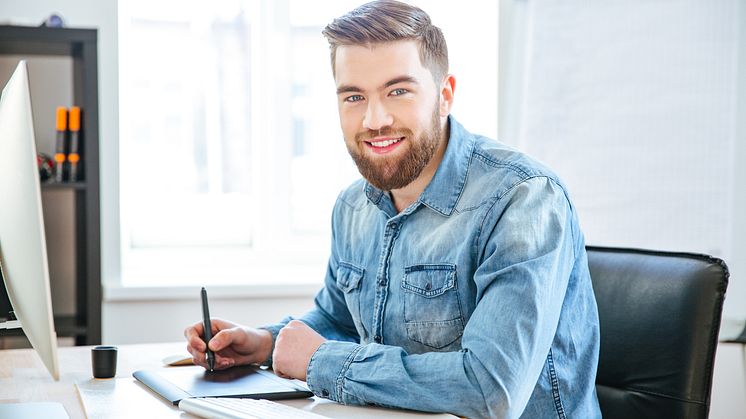 Junger Mann mit Bart sitzt lächelnd am Schreibtisch, arbeitet mit einem Grafiktablet und trägt ein Jeanshemd.