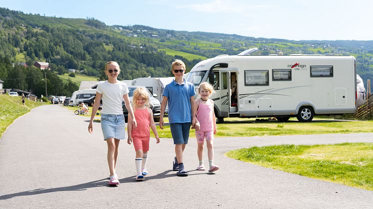 Fire barn går hånd i hånd på en campingplass en solfylt sommerdag, med bobiler og grønne åser i bakgrunnen