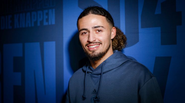 Taylan Bulut steht vor einer blauen Wand mit S04-Schriftzug.