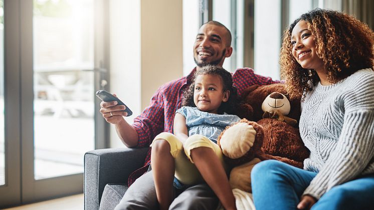 Lächelnde Familie sitzt gemeinsam mit Kind und Teddybär auf dem Sofa und schaut fern.