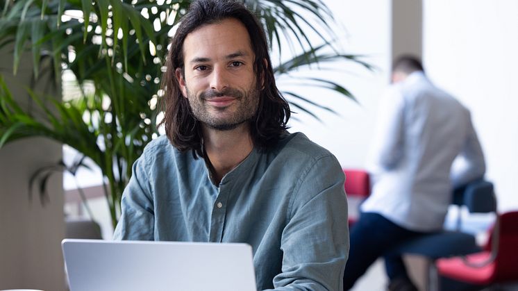 Mann mit langen Haaren sitzt lächelnd am Laptop in einem modernen Büro mit Pflanzen im Hintergrund.