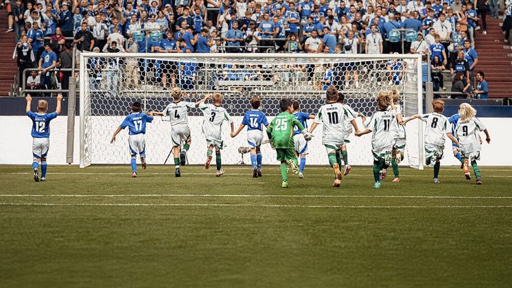 Eine Gruppe von jungen Fußballern rennen auf ein großes Tor zu, während viele Zuschauer auf einer Tribüne zuschauen