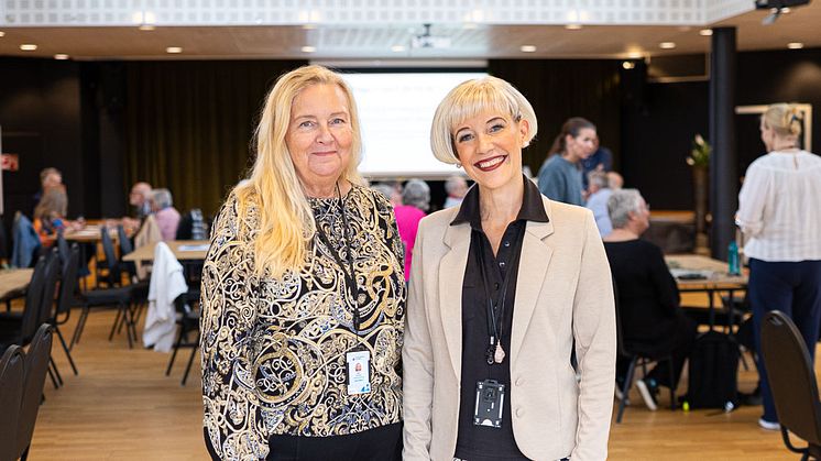 Två kvinnor poserar leendes i en lokal där många sittande och stående workshopdeltagare skymtas i bakgrunden.