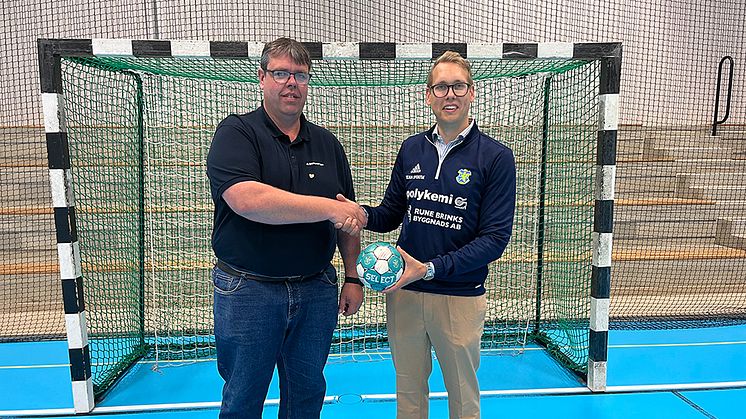 Två män, varav den ena håller en handboll, skakar hand framför ett handbollsmål i en idrottshall