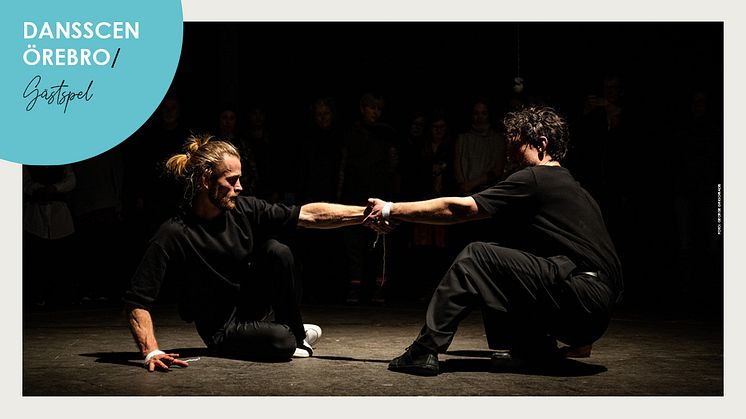 Två manliga dansare sitter på scengolv och håller i varandras utsträckta arm.
