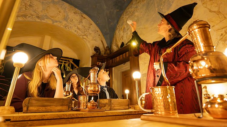 eine Hexenmeisterin und drei Hexenlehrlinge - sie schauen interessiert zu, welchen Stein die Hexenmeisterin in ihrerHand hält