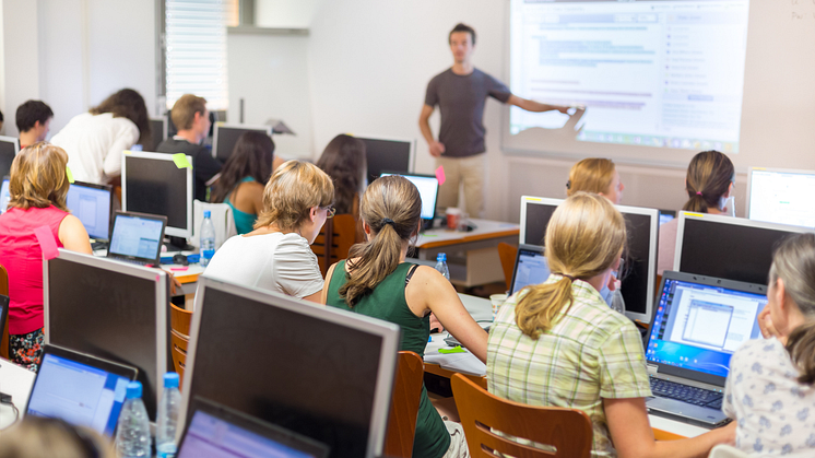 Image of a group of people sat at desks with computers looking at a man teaching infront of a projector screen. 