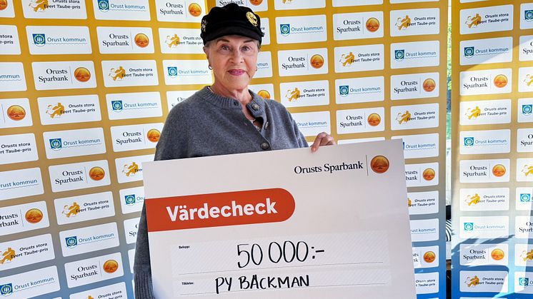 Py Bäckamn håller en stor symbolisk check på 50 000 kronor framför en fotovägg med Orusts Sparbank och Orusts kommun.