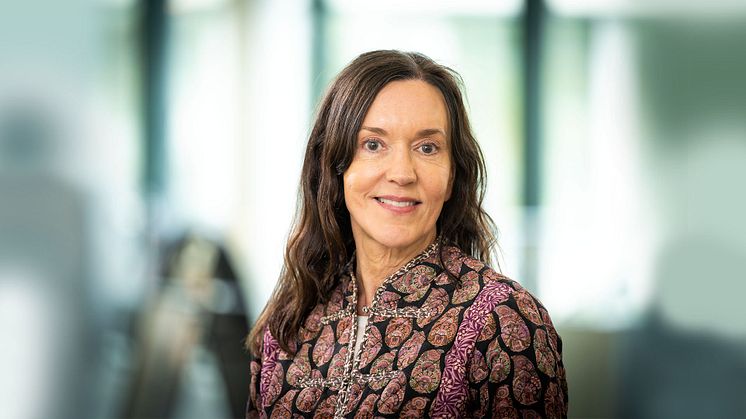 Susanna Toivanen tittar in i kameran. Hon har långt, brunt hår och en lilamönstrad skjorta på sig. Bakgrunden är suddig.