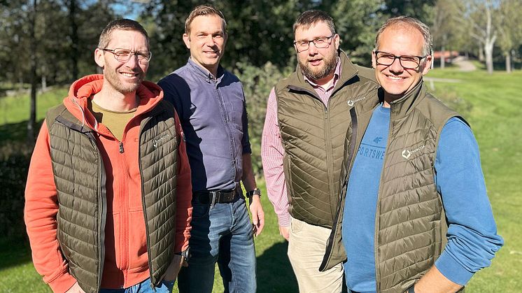 Bilden är ett gruppfoto på fyra personer som står utomhus i en parkliknande miljö.