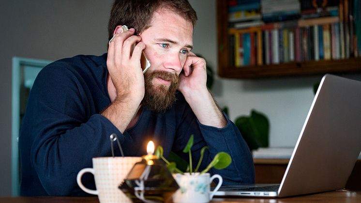 En man med skägg sitter vid ett bord i hemmiljö framför en dator och pratar i telefon. På bordet står ett ljus och en tekopp.