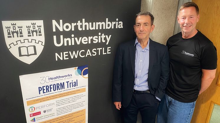 The image shows two men standing next to each other against a black wall with a white Northumbria University logo on it