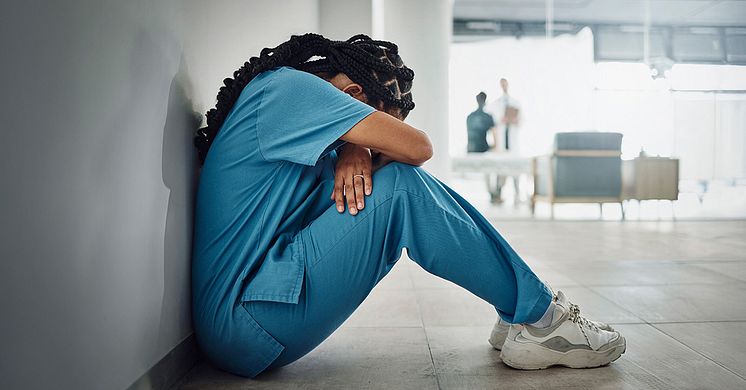 En undersköterska sitter på golvet med ryggen mot en vägg, hon lutar sitt huvud mot i armarna som hon håller om sina knän. 