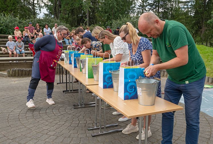 En rekke med mennesker som står ved et langbord og skreller poteter i hver sin bøtte.