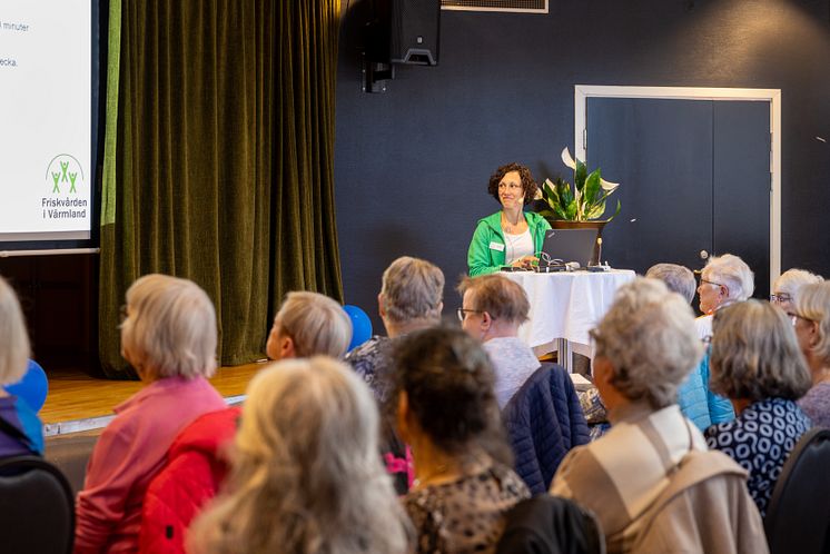 a.	Talare med öronmikrofon står framför en stor sittande publik av seniorer. Hennes blick är vänd mot presentationen som visa