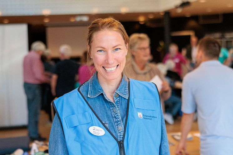 Leende kvinna i blå funktionärsväst står i mässhall, med många besökare som syns bakom henne.