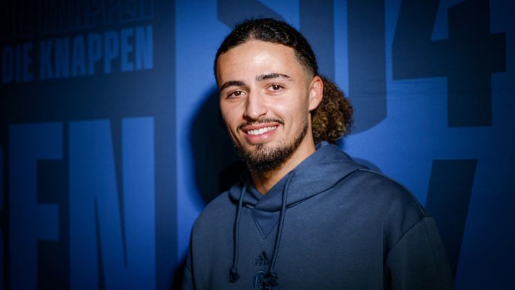 Taylan Bulut steht vor einer blauen Wand mit S04-Schriftzug.