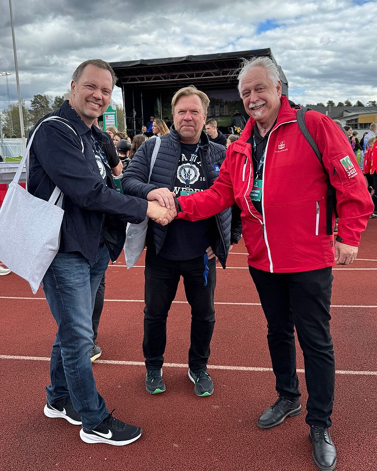 Tre personer på en idrottsarena skakar hand. 