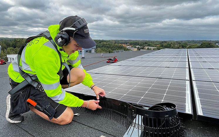 Montör från Nordic Solar monterar solceller på ett stort fastighetstak, med  säkerhetssele under klar himmel.