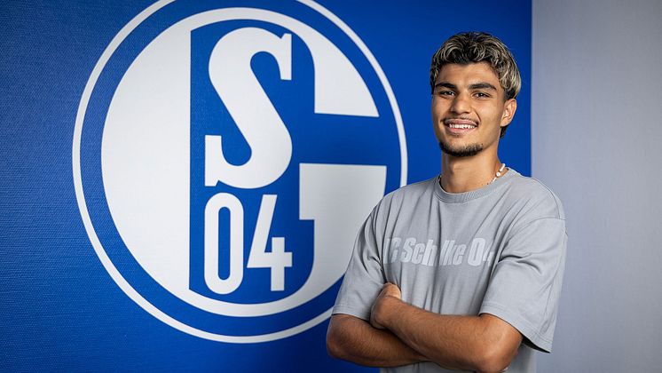 Mertcan Ayhan steht vor einer Wand mit Schalke Logo