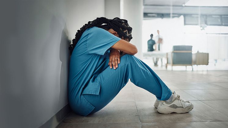En undersköterska sitter på golvet med ryggen mot en vägg, hon lutar sitt huvud mot i armarna som hon håller om sina knän. 
